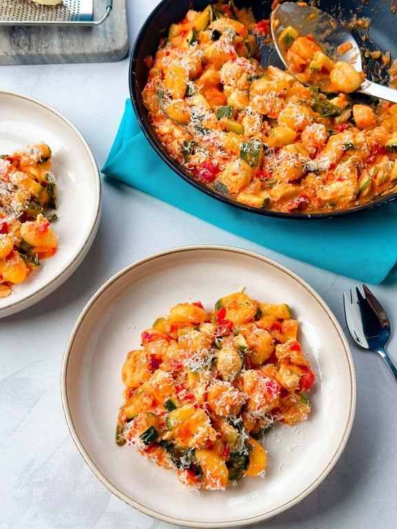 Two plates and a pan of gnocchi with vegetables and grated cheese on a blue napkin