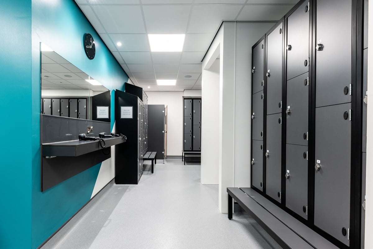 PureGym Consett: Locker room with benches, sinks, multiple enclosed shower stalls.