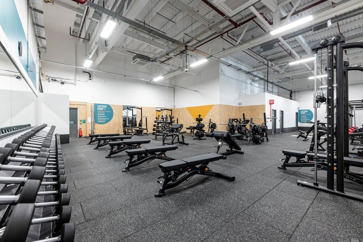 PureGym Rushden: Strength training area with benches, squat racks, free weights, and rubberized flooring.
