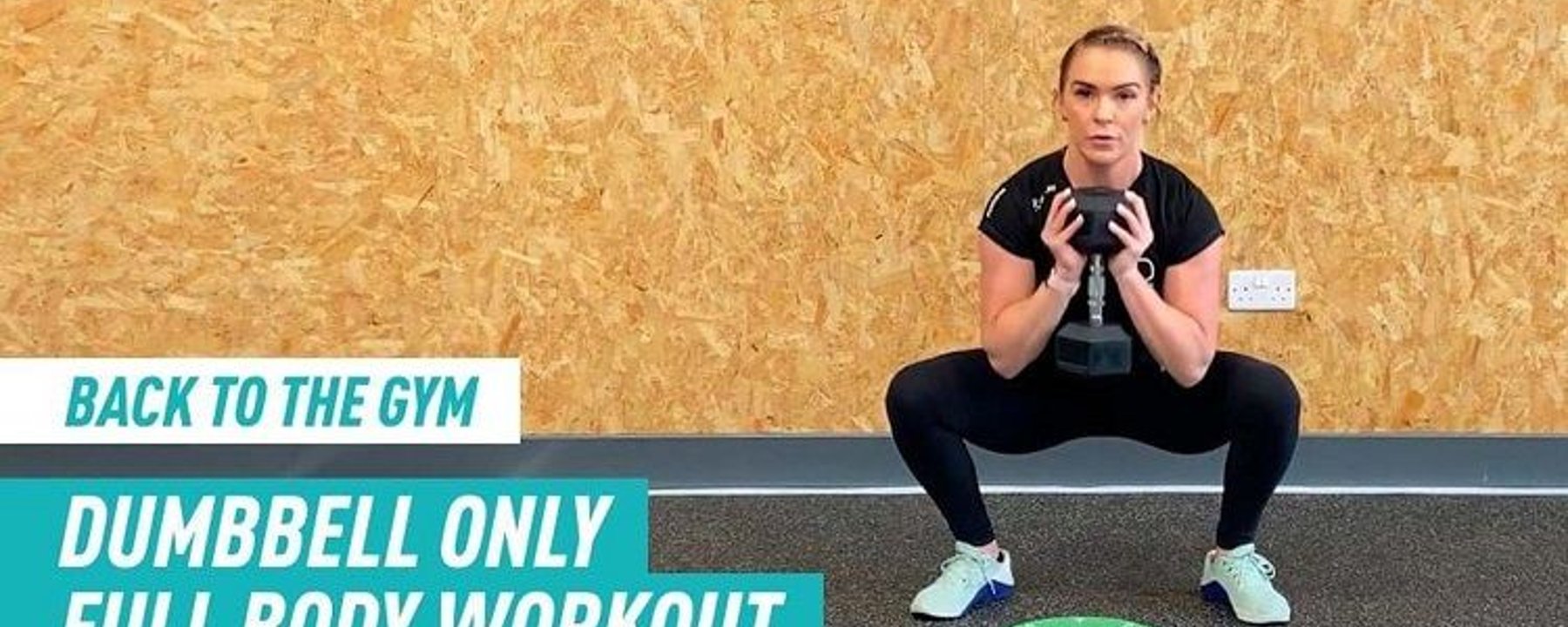 Woman in black workout clothes holding dumbbell in a squat position on gym floor