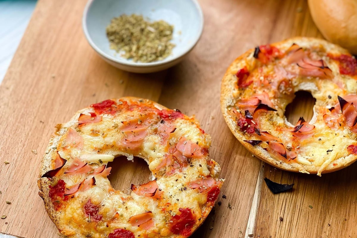 Two toasted bagel halves with melted cheese and ham on a wooden board with herbs nearby