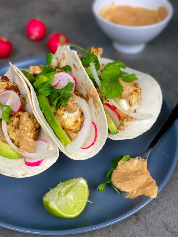 Three tacos with cauliflower, radish, onion, avocado, coriander, and sauce on a blue plate