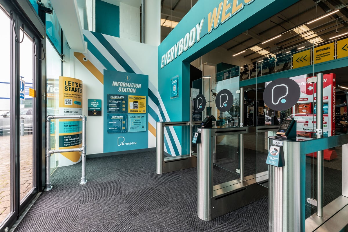 PureGym Caerphilly: Reception area with information signage, self-service kiosks, turnstiles.