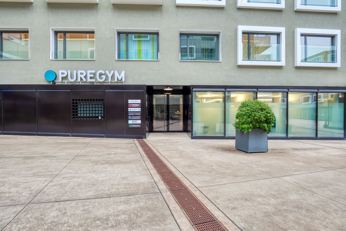 Der Ausseneingang eines PureGym mit Beschilderung und Glastüren, gelegen in einem gepflasterten Aussenbereich
