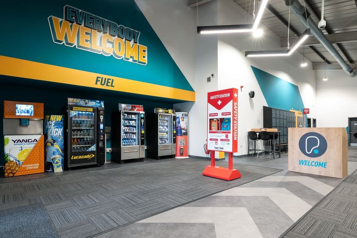 PureGym Leeds Bramley: Beverage vending coolers, signage that says "Everybody Welcome," and a kiosk.