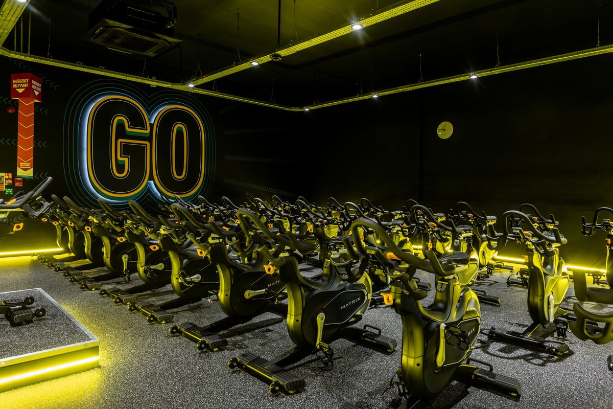 PureGym Peterborough Brotherhood Retail Park: Rows of stationary bikes in a dimly lit studio with neon "GO" sign illuminated.