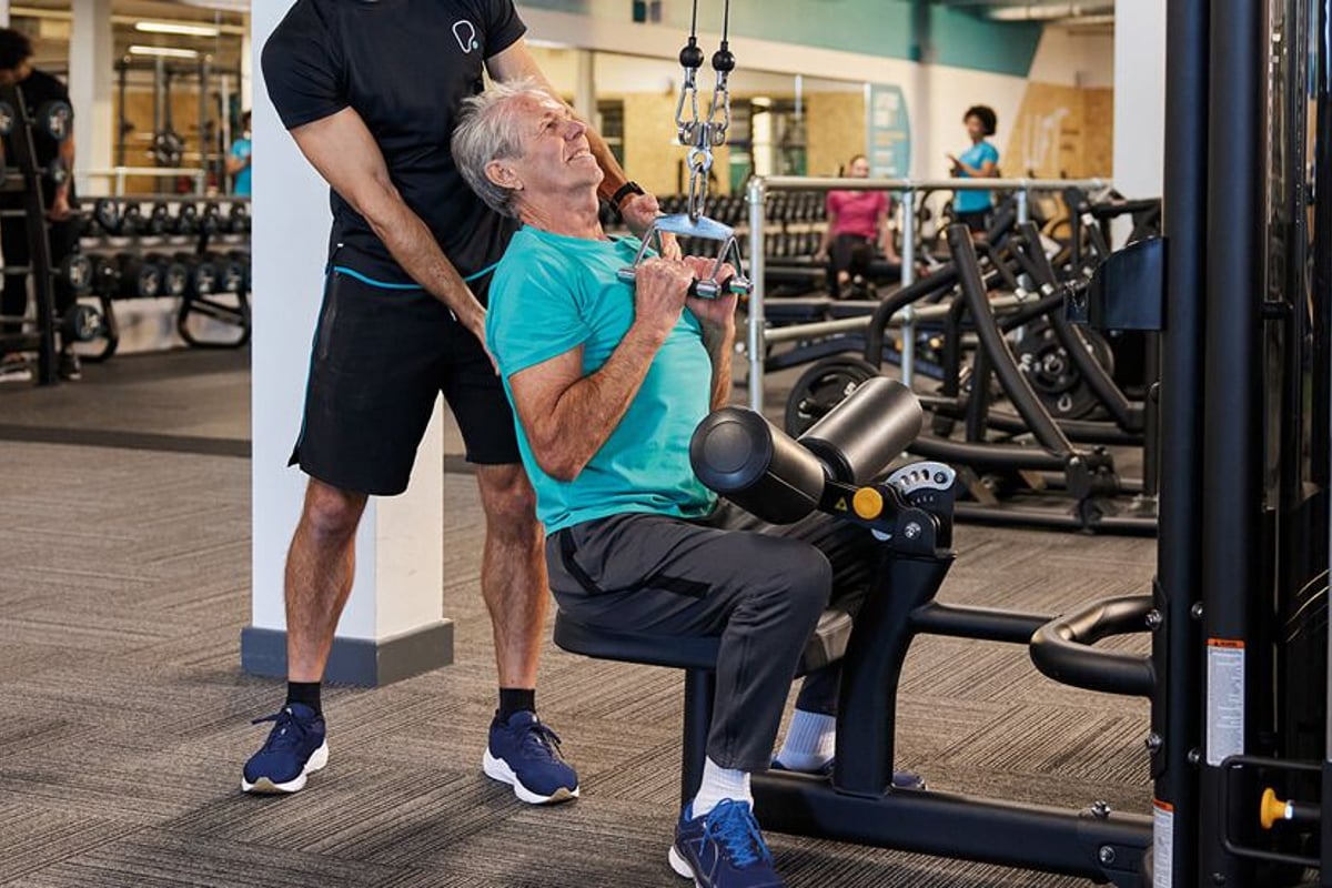 Older man doing lat pulldown exercise with trainer assistance in gym