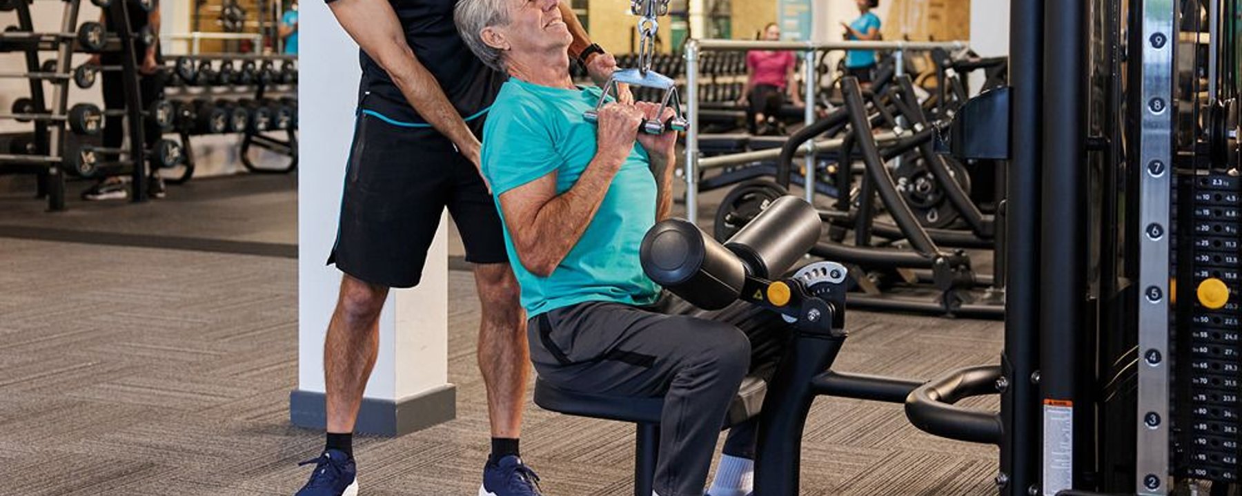 Older man doing lat pulldown exercise with trainer assistance in gym