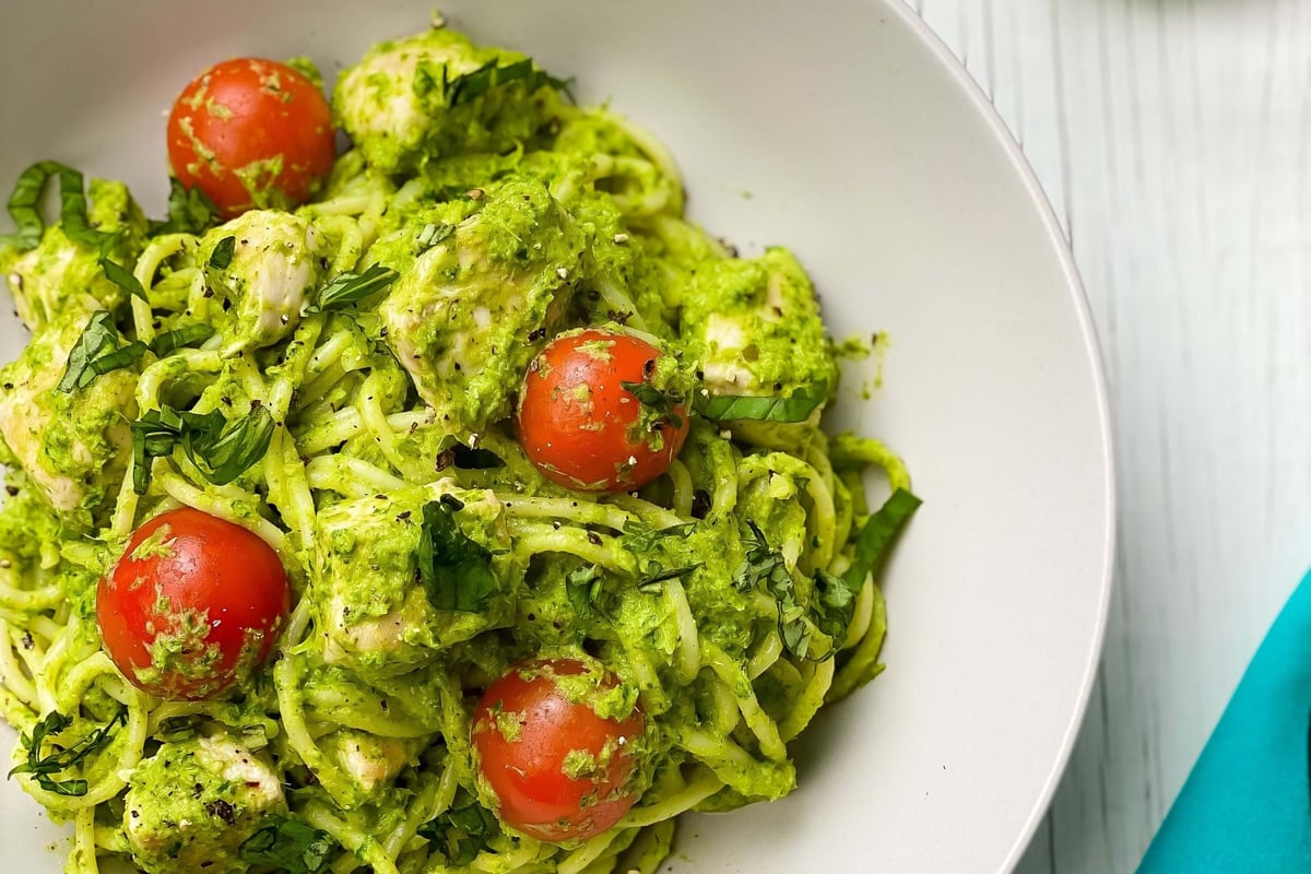 Pasta with green pesto sauce, cherry tomatoes, and fresh herbs in a white bowl.