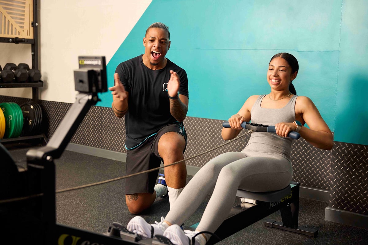 Woman rowing on machine while trainer encourages her in gym with teal wall background