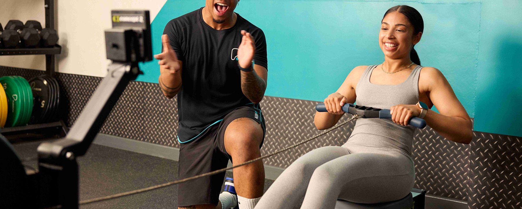 Woman rowing on machine while trainer encourages her in gym with teal wall background