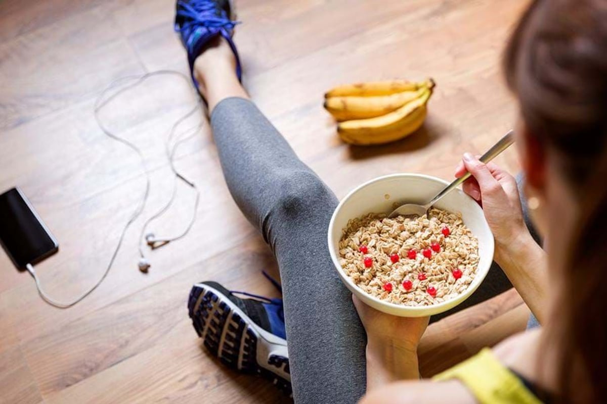 Person in sportswear sitting on wooden floor eating cereal with red berries and bananas ne