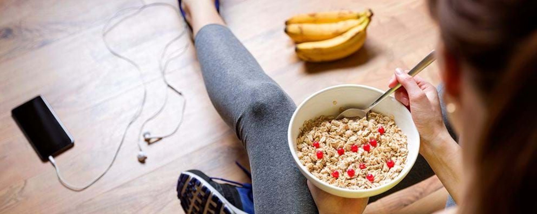 Person in sportswear sitting on wooden floor eating cereal with red berries and bananas ne