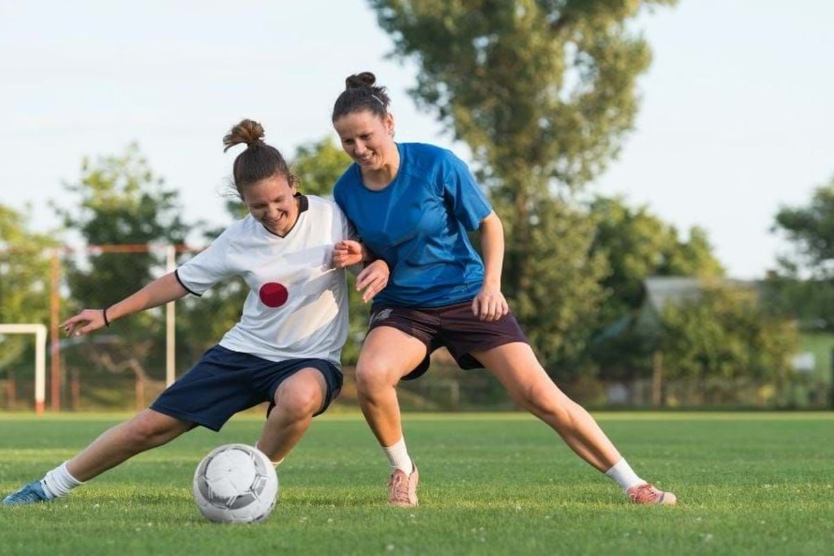 Two women competing for a football on a grassy pitch with trees in the background