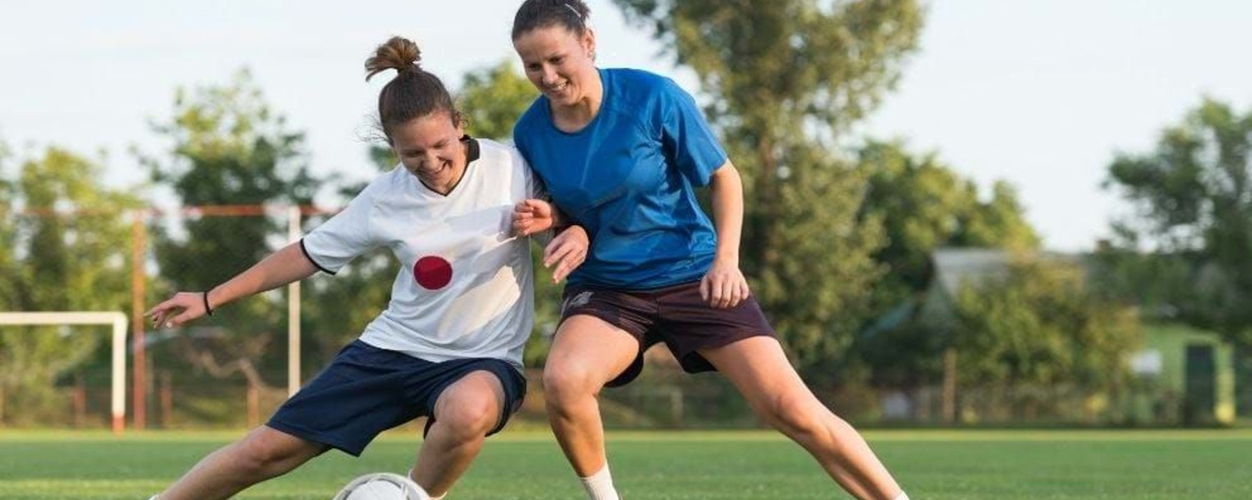 Two women competing for a football on a grassy pitch with trees in the background