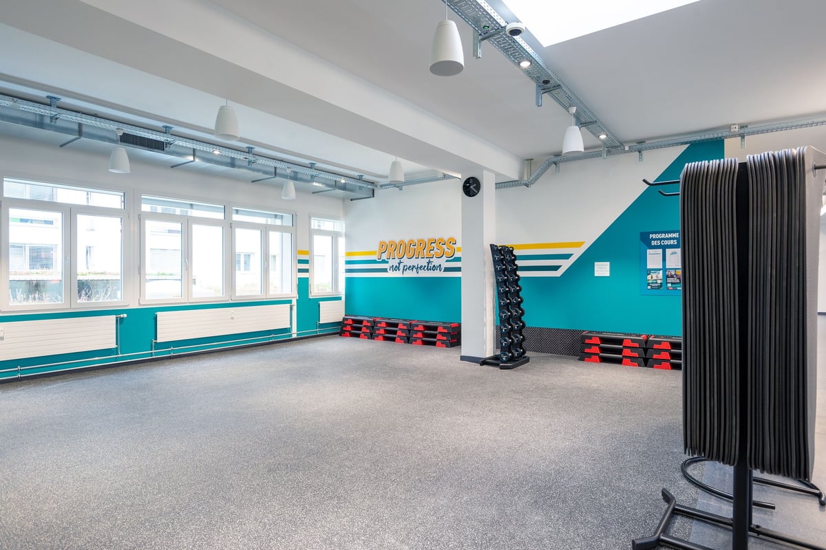 PureGym Vevey: Murs bleu sarcelle, compartiments de rangement, barres murales, bandes de résistance et sols caoutchoutés dans un studio de fitness.