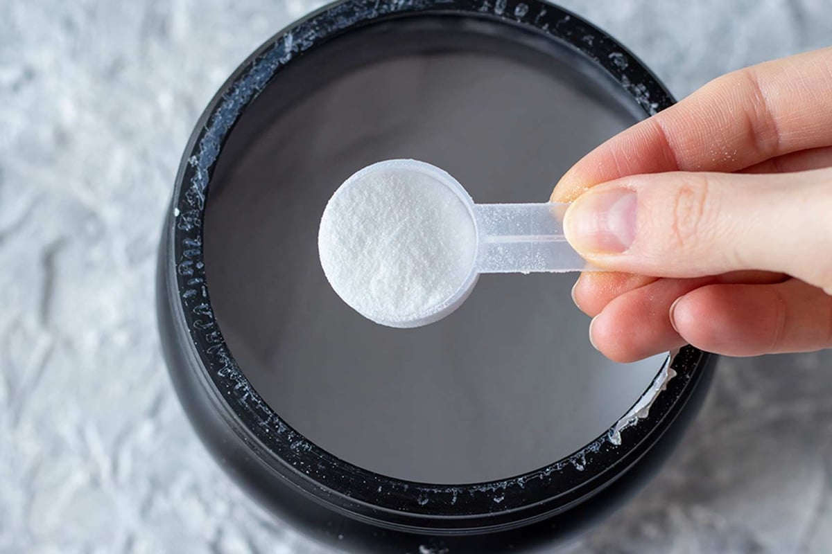 Hand hält Messlöffel mit weißem Pulver über dunkler Flüssigkeit in Schüssel.