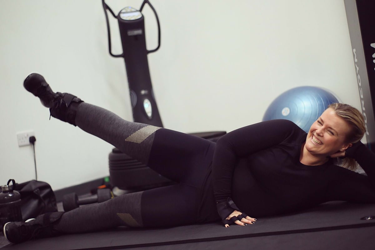 Woman in black sportswear lying on side lifting leg on gym mat with exercise ball nearby
