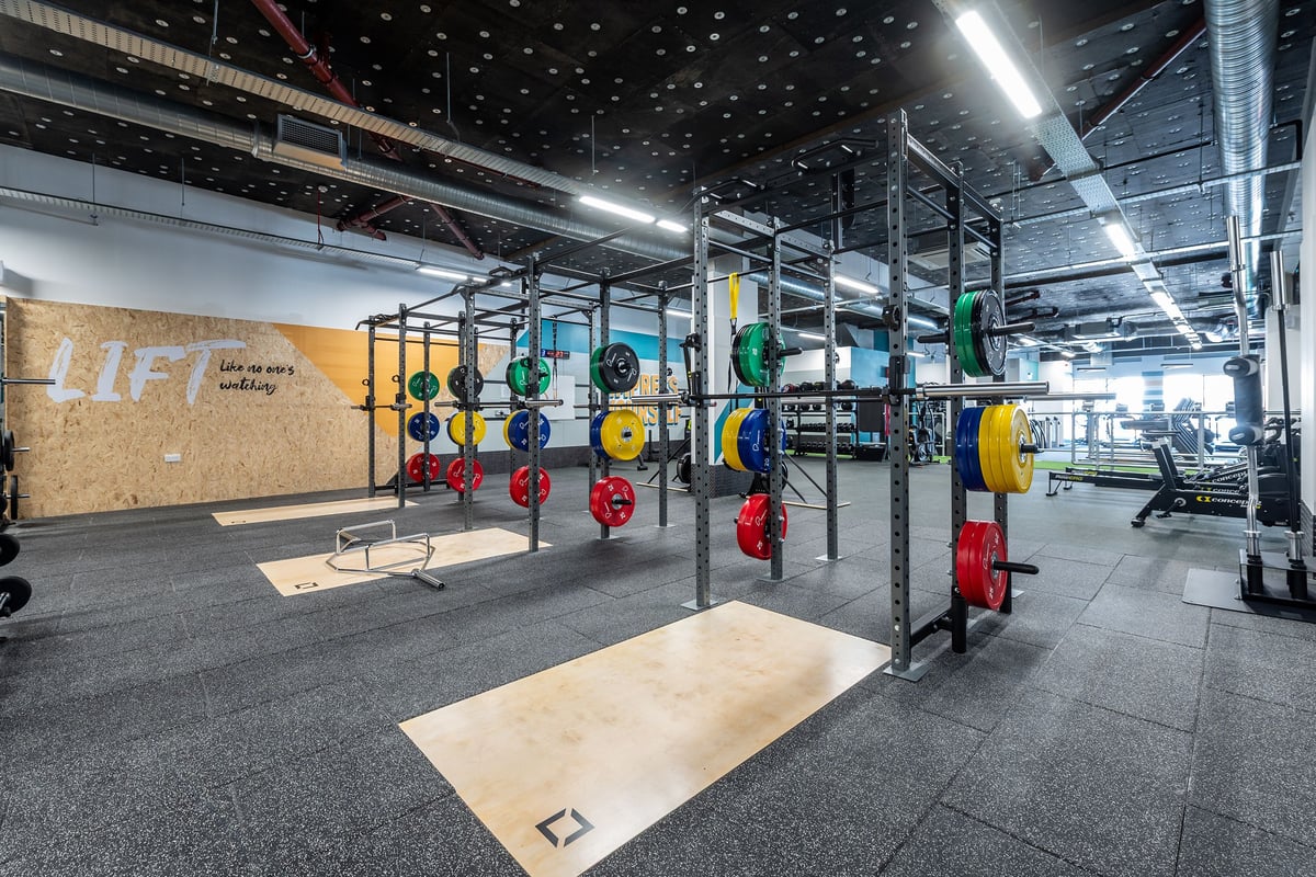 PureGym London Beckenham: Rig of weighted bars, colourful weight plates, rubberised flooring in a dimly lit gym area.