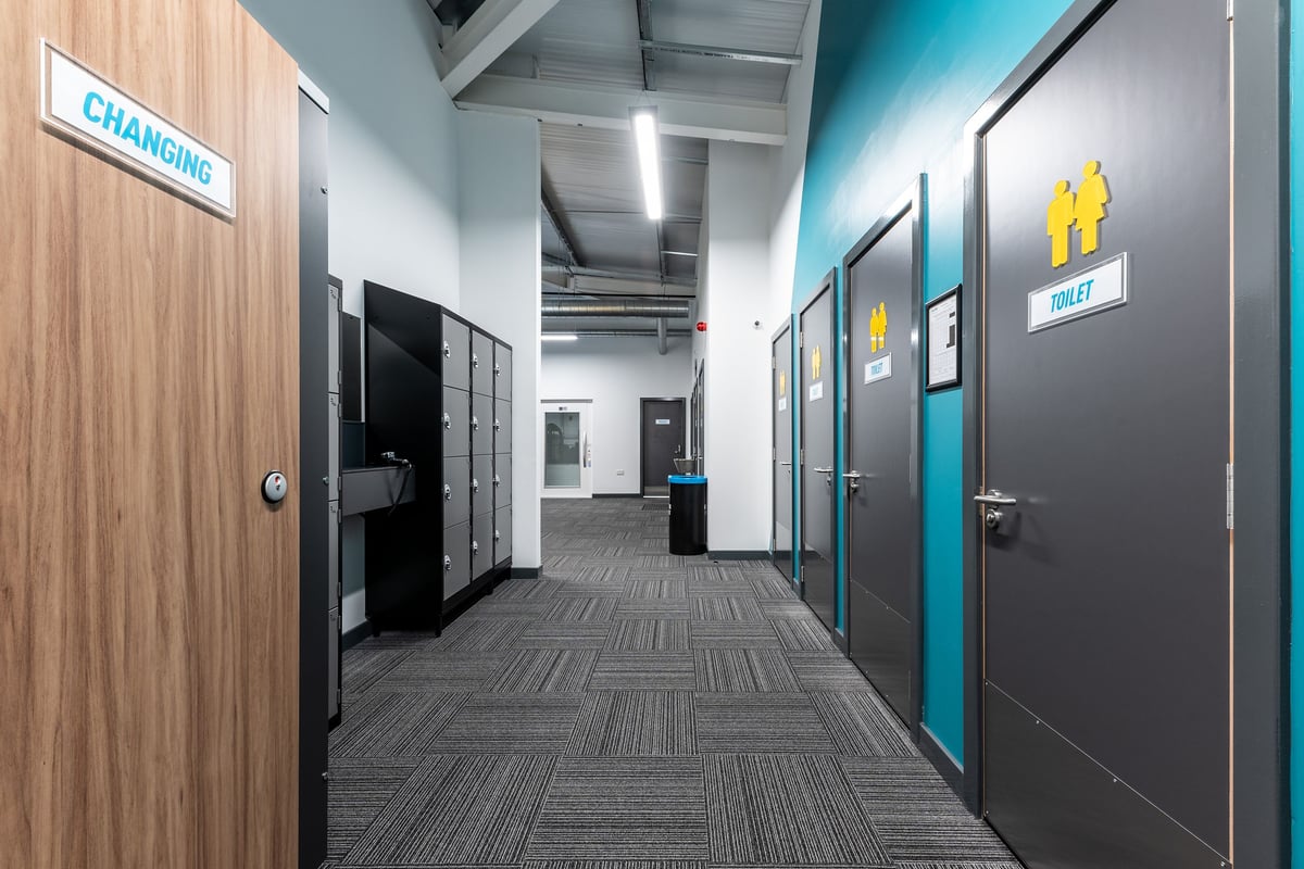 PureGym Galashiels: Changing room corridor with cubicles, carpeted floor, signage indicating facilities.