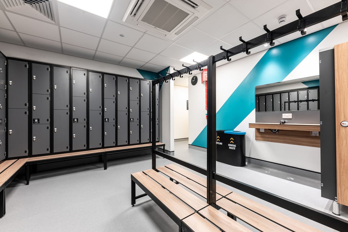 PureGym Liverpool Edge Lane: Rows of lockers, wooden benches, and entry turnstiles in a locker room setting.