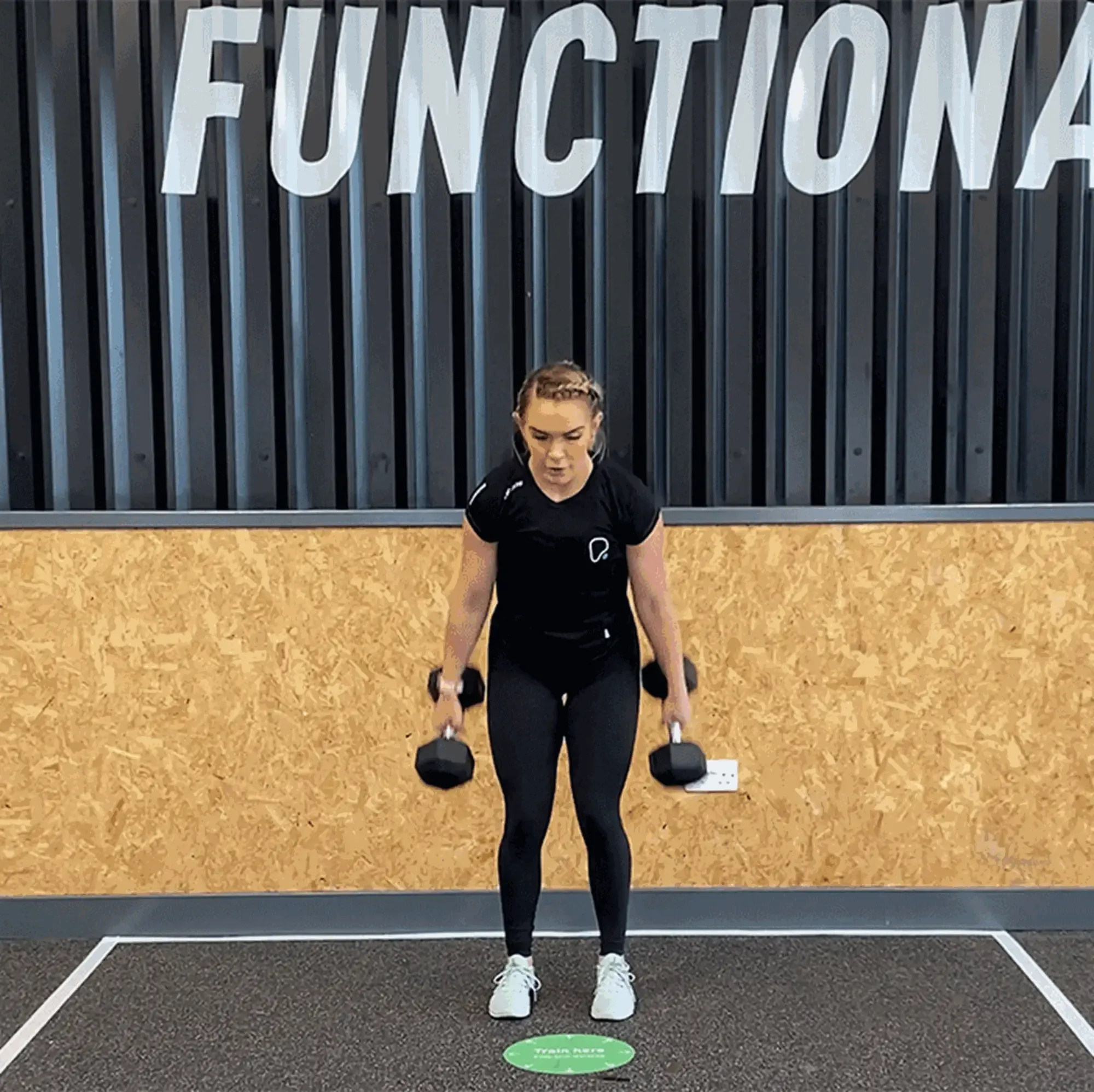 Woman in black workout clothes lifting dumbbells in gym with FUNCTIONAL sign above
