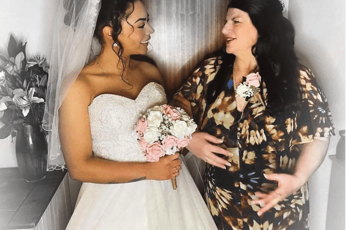 Bride in white dress holding bouquet talking to woman in patterned dress indoors