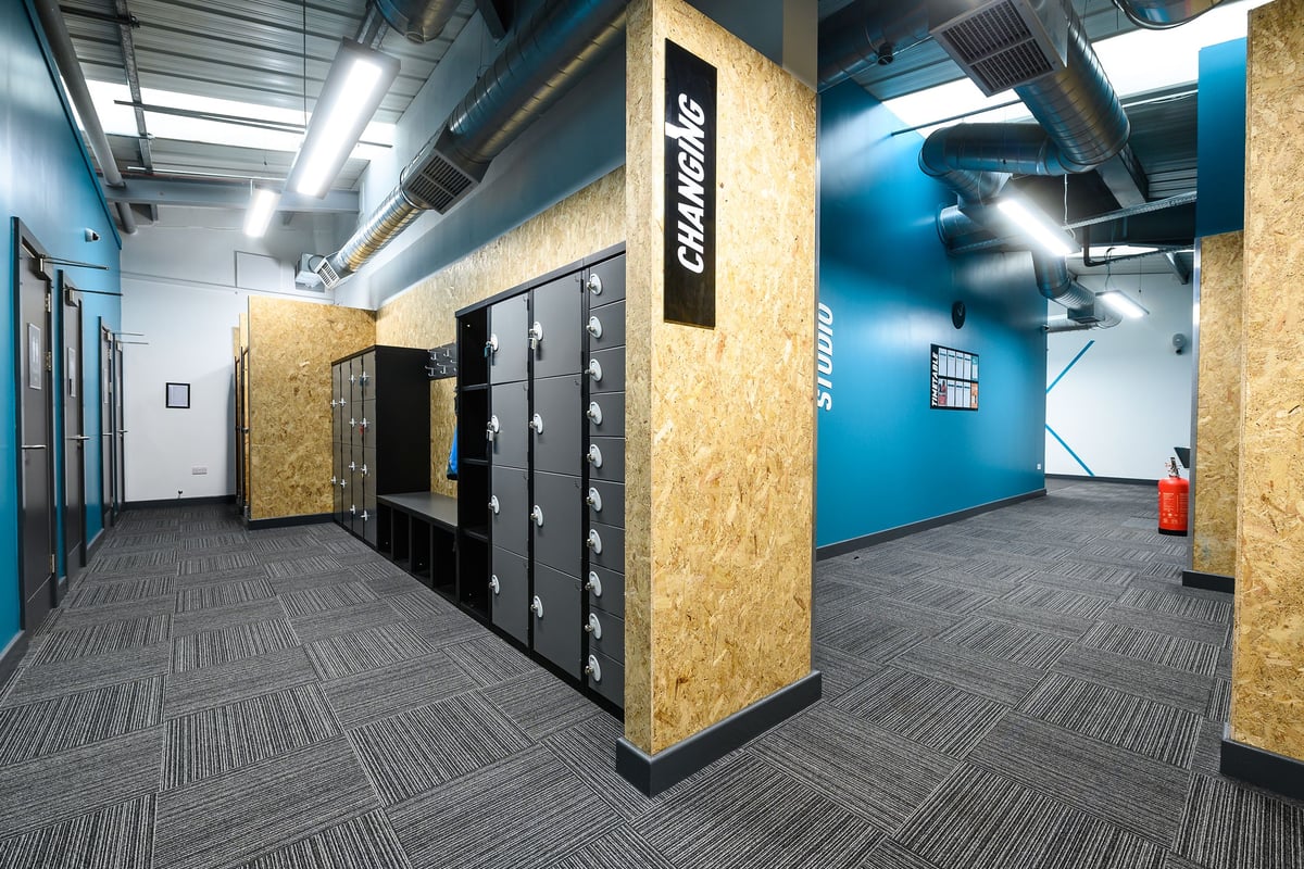 PureGym Glossop: Lockers, ventilation ducts, carpeted floor in a dimly lit, industrial-style interior space.