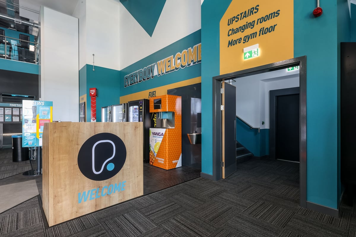 PureGym Bishop Auckland: Gym entrance with signage, self-service kiosk, and cardio area visible upstairs.
