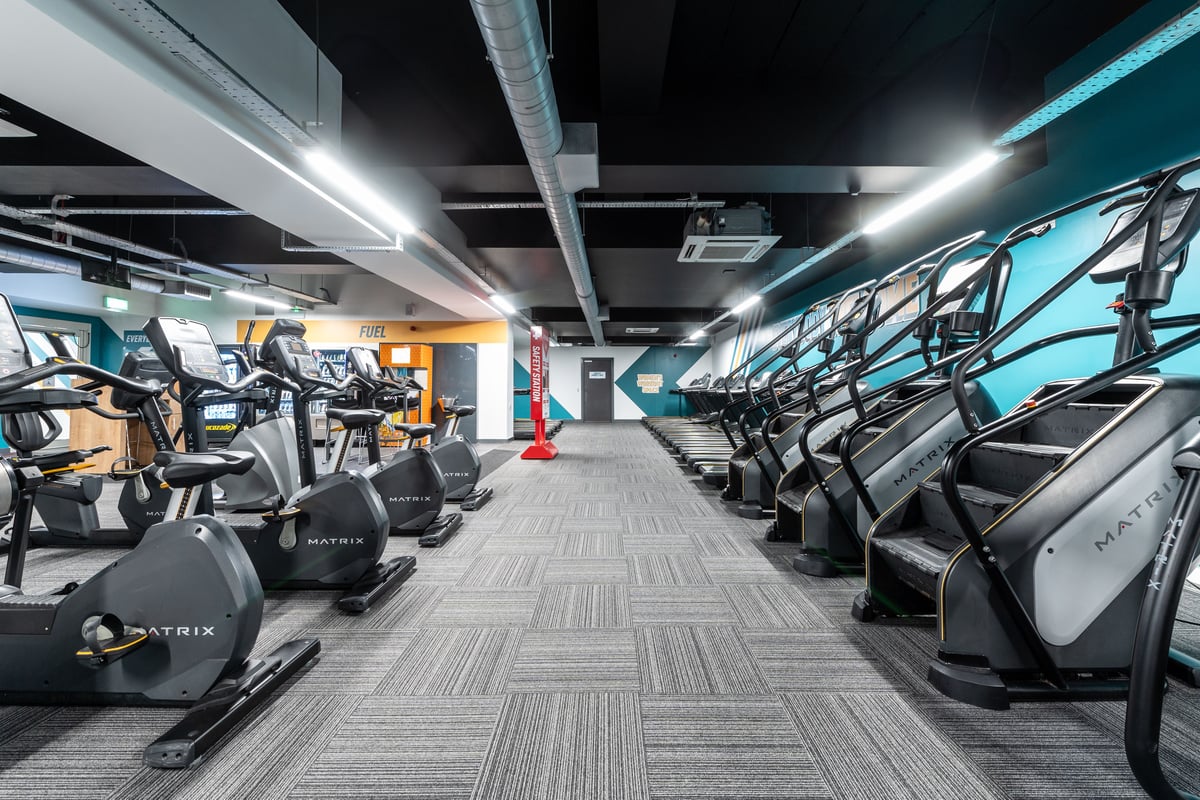 PureGym London Ilford: Rows of elliptical machines and treadmills in a dimly lit gymnasium with blue accent lighting.