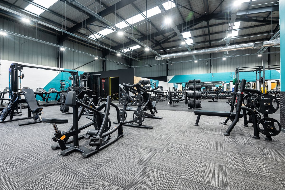 PureGym Dunfermline: Rows of seated weightlifting machines, free weights, and rubberized flooring in a spacious gym area.