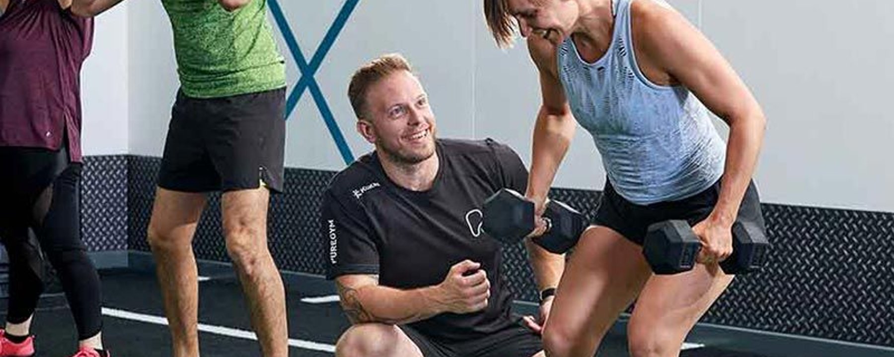 Man coaching woman lifting dumbbells in gym while others exercise nearby