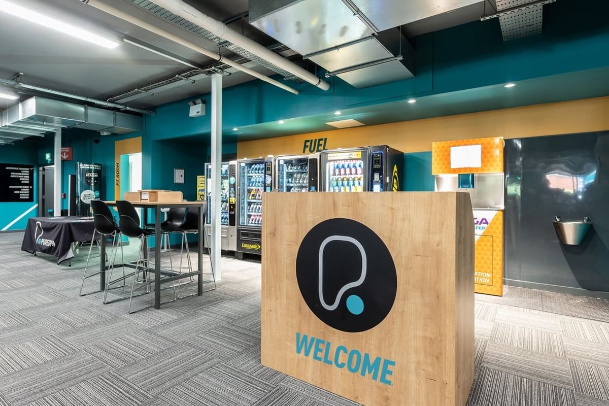 PureGym Oldham: Welcome desk, vending machines, bar stools in a modern cafÃ© or lounge area.