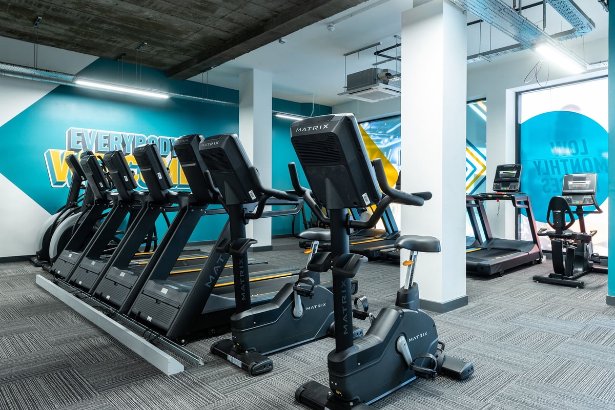PureGym London Streatham: Rows of treadmills, exercise bikes, and ellipticals in a bright teal and yellow gym studio.