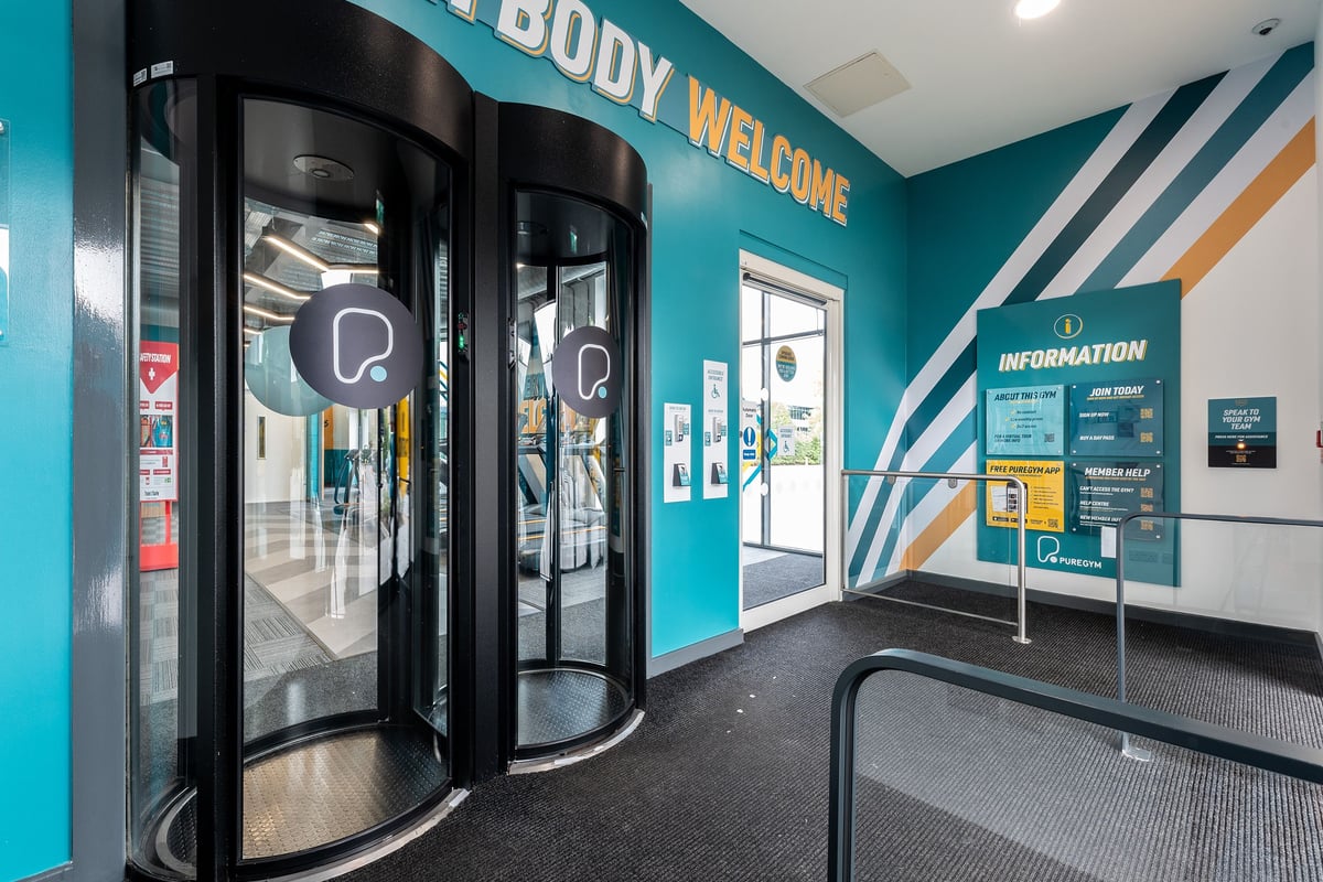 PureGym Reading Basingstoke Road: Entrance area with information stands, glass doors, and teal branding for a fitness centre.
