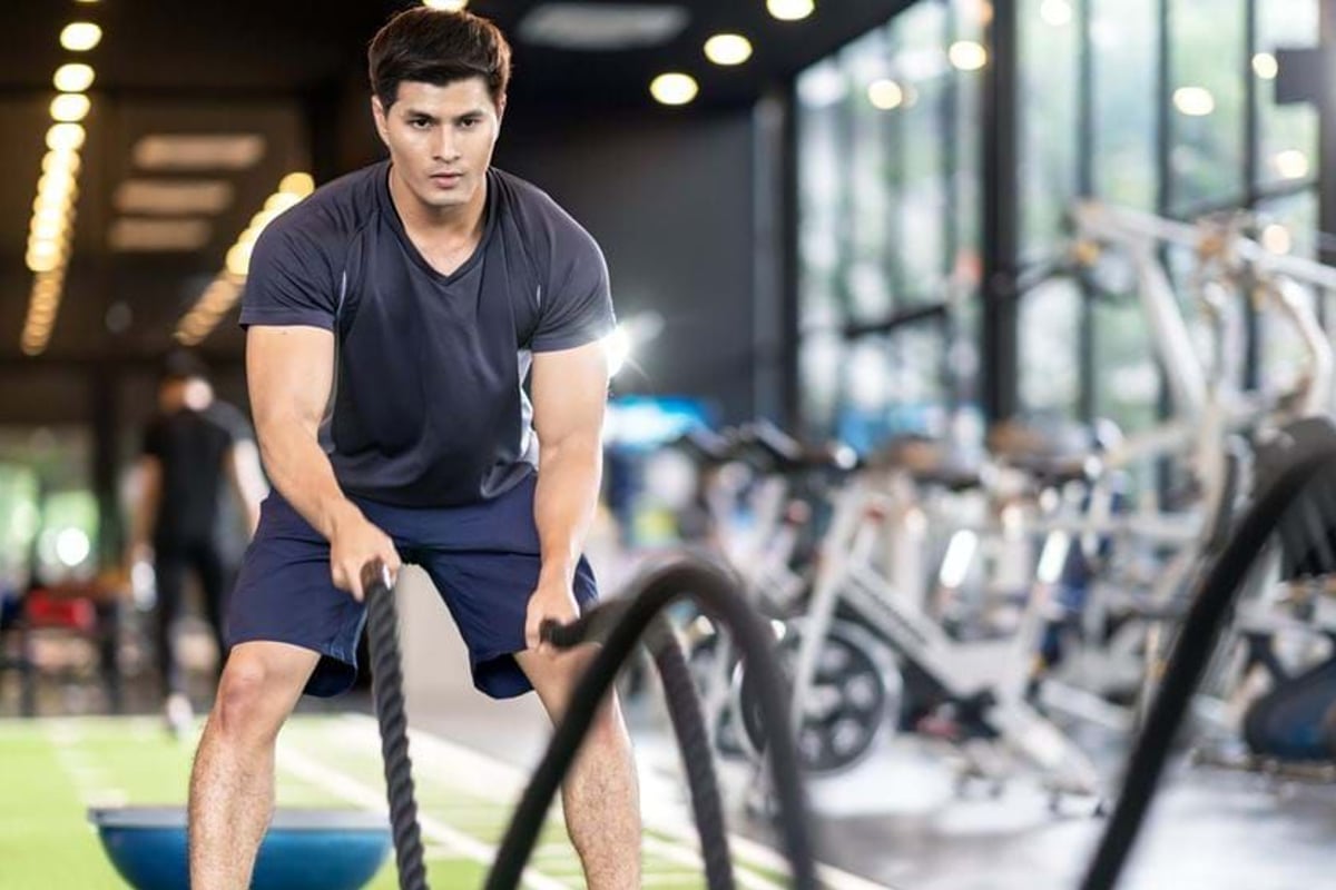 Man exercising with battle ropes in a gym with exercise bikes in the background