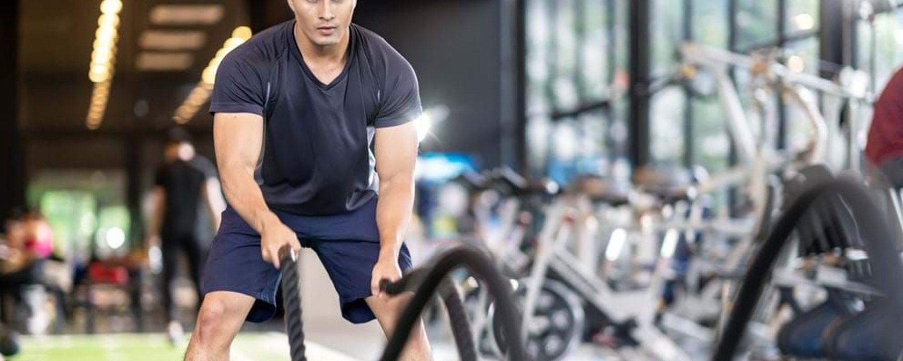 Man exercising with battle ropes in a gym with exercise bikes in the background
