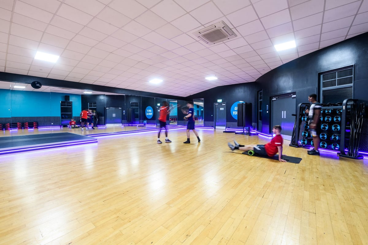 PureGym Manchester Moston: Mirrored studio with boxing bags, weight racks, wooden floors, and purple lighting.
