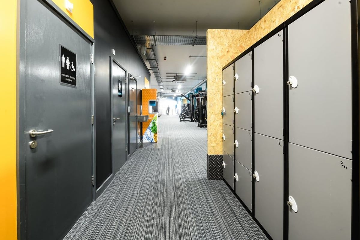 PureGym East Grinstead: Corridor with grey lockers, tiled flooring, and wall-mounted lamps.