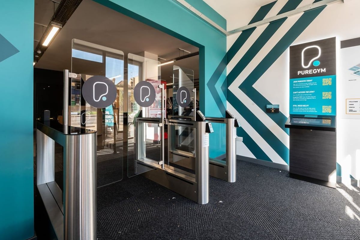 PureGym London Catford Rushey Green: Entry gates with full-length glass doors and modern signage on teal walls.