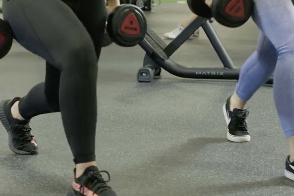 Two people lunging with dumbbells in a gym with workout equipment in the background