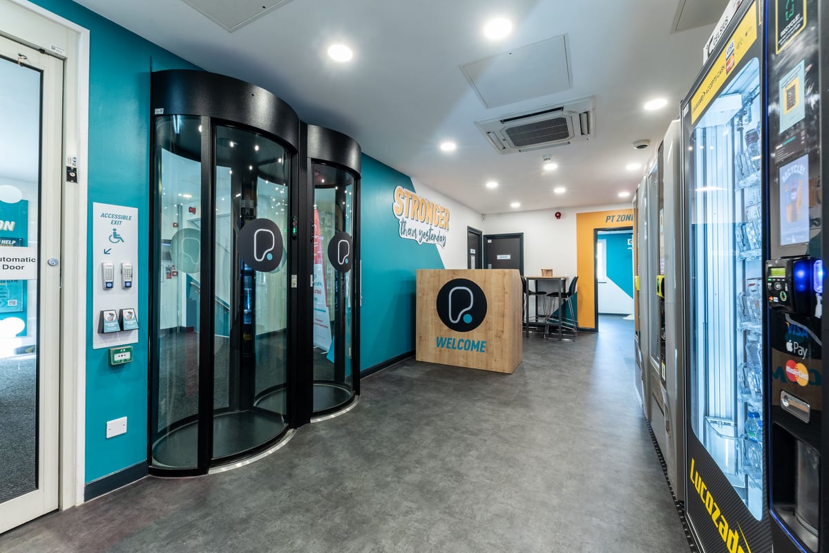PureGym London Muswell Hill: Revolving doors leading into a corridor with self-service kiosks and teal-coloured walls.