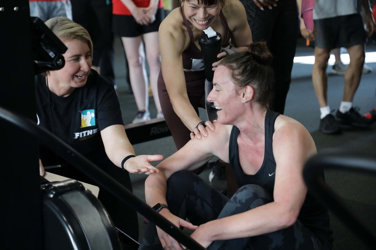 Three women smiling and interacting near a rowing machine in a gym setting