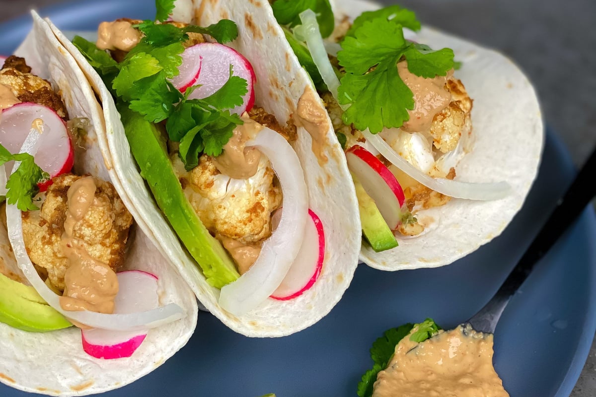 Three tacos with cauliflower, radish, onion, avocado, coriander, and sauce on a blue plate
