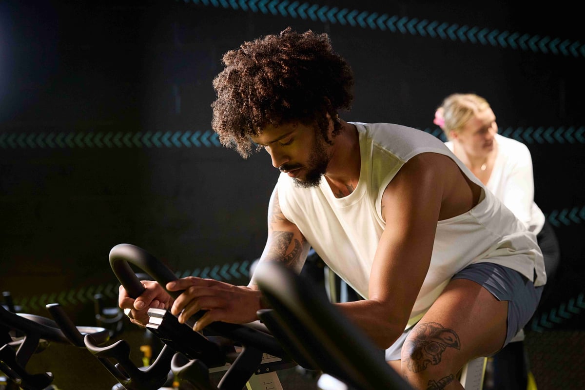 Man with tattoos cycling on exercise bike in dark gym with woman behind him