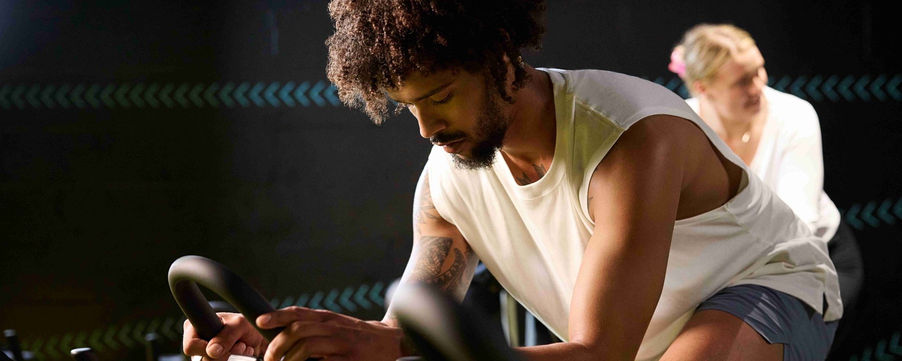 Man with tattoos cycling on exercise bike in dark gym with woman behind him