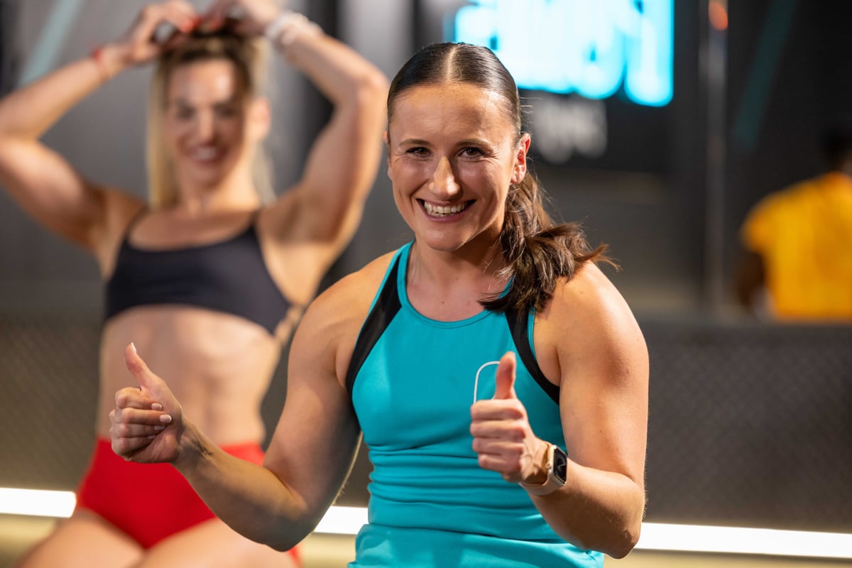 Smiling woman in blue sportswear showing thumbs up with another woman in background.