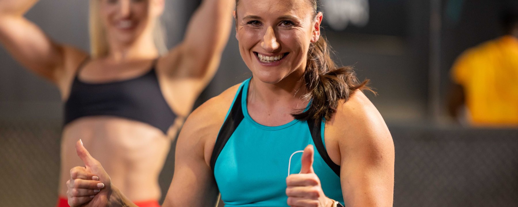 Smiling woman in blue sportswear showing thumbs up with another woman in background.