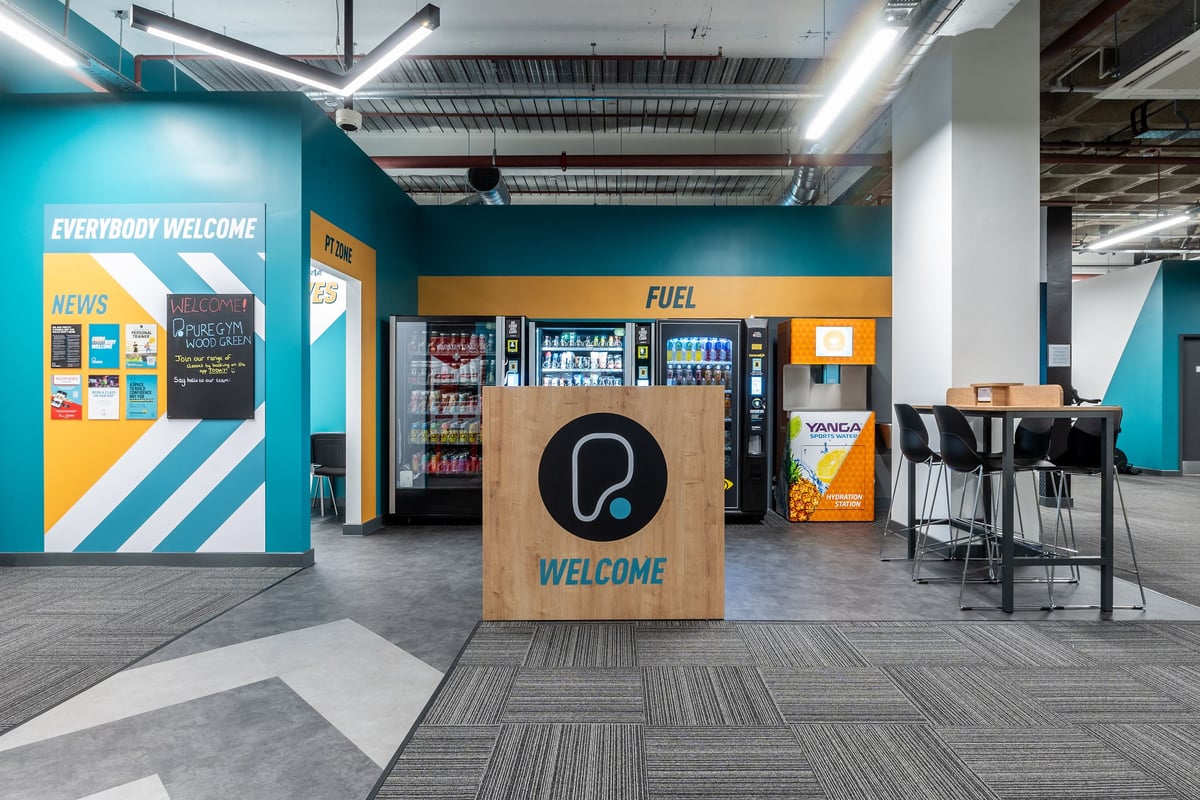 PureGym London Wood Green: Vending machines, cafÃ© seating area, and welcoming signage in brightly coloured interior space.