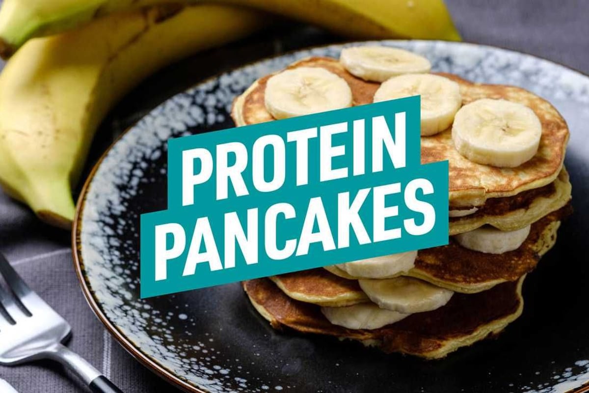 Stapel Pfannkuchen mit Bananenscheiben auf schwarzem Teller, daneben Banane und Gabel